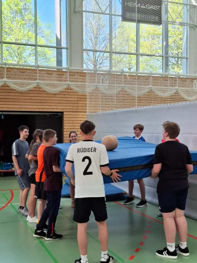 Mehrere Jugendliche stehen in einer Turnhalle. Sie heben gemeinsam eine blaue Matte, auf der ein großer Ball liegt. Im Hintergrund sind hohe Fenster mit Blick auf grüne Bäume zu sehen.