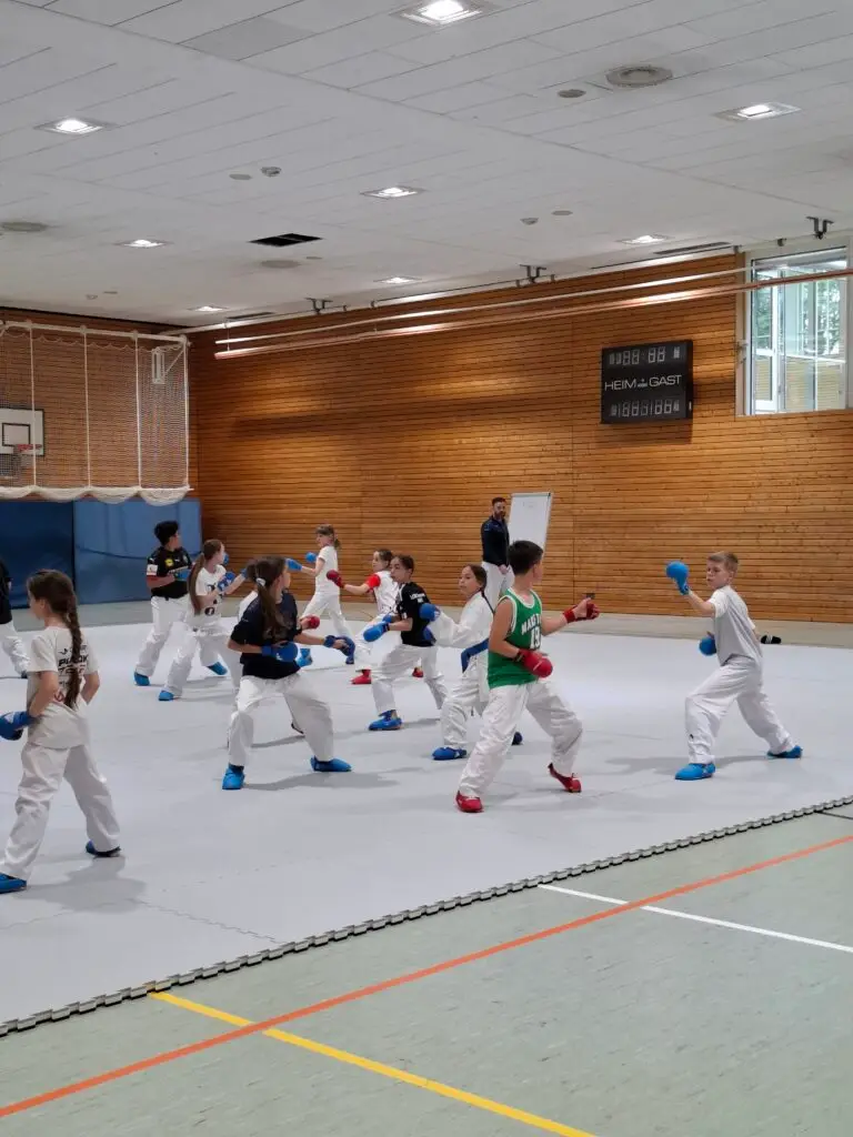 Kinder trainieren Karate in einer Turnhalle, tragen weiße Anzüge und bunte Schutzhandschuhe. Ein Trainer beobachtet im Hintergrund. Auf einer Anzeigetafel steht "Heim:Gast". Holzverkleidung und Sportmatten sind sichtbar.