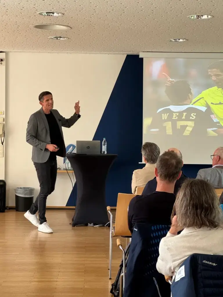 Ein Mann hält einen Vortrag vor einem sitzenden Publikum. Er steht neben einem Notebook auf einem Stehpult und zeigt auf die Leinwand, auf der ein Bild von einem Fußballschiedsrichter und einem Spieler mit der Rückennummer 17 zu sehen ist. Der Vortrag findet in einem modernen Raum mit hölzernem Boden statt.