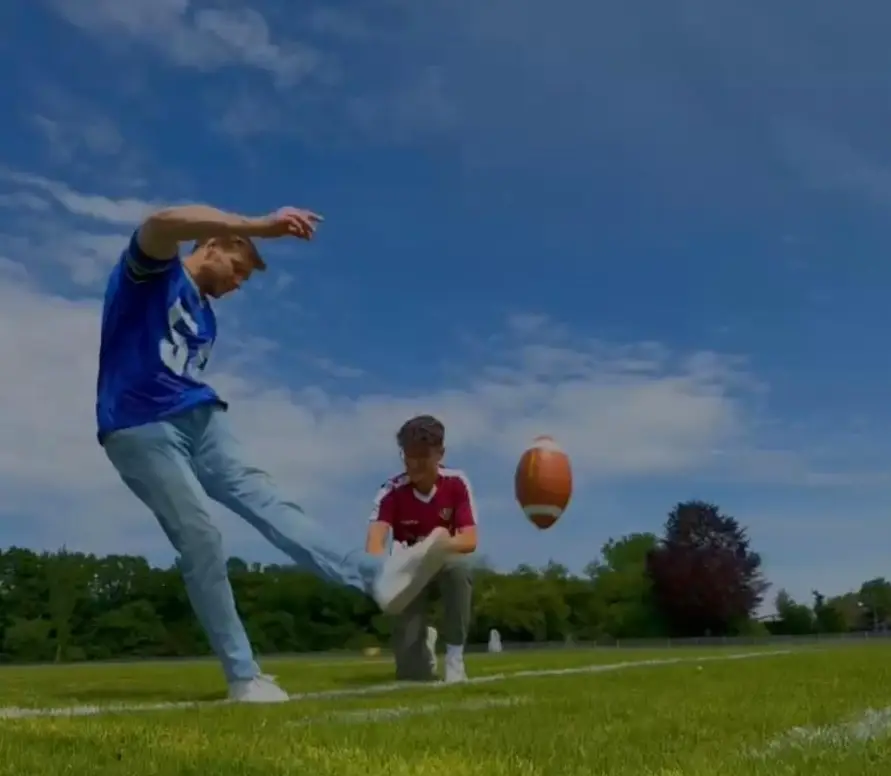 Ein Mann in einem blauen Trikot schießt einen Football, der von einem zweiten Mann im roten Trikot gehalten wird, auf einem grünen Sportfeld. Der Himmel ist leicht bewölkt.