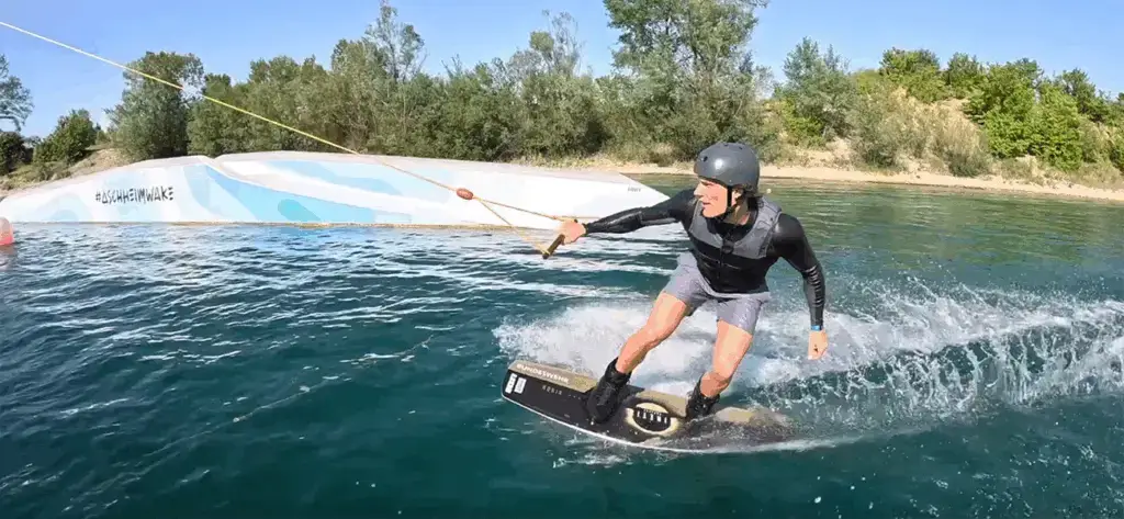 Person beim Wakeboarden auf einem See, trägt Helm und Neoprenanzug, hält an einem Seil fest. Im Hintergrund ist ein Hindernis mit der Aufschrift "#ascheimwake" und Bäume zu sehen. Der Himmel ist klar und blau.