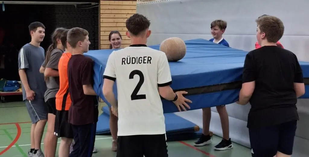 Mehrere Jugendliche halten zusammen eine Turnmatte in einer Sporthalle, während ein Medizinball darauf liegt. Einige der Jugendlichen lächeln.
