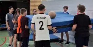 Mehrere Jugendliche halten zusammen eine Turnmatte in einer Sporthalle, während ein Medizinball darauf liegt. Einige der Jugendlichen lächeln.