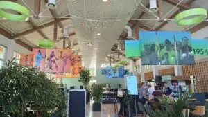 Ein öffentlicher Bereich mit hohen Decken und viel Tageslicht. Farbige Banner mit Sportbildern und historischem Fotomaterial hängen von der Decke. Innenbegrünung mit großen Pflanzen dominiert den Raum. Rechts befindet sich ein Café mit Menschen, die sich unterhalten. Ein Schild listet Speisen und Getränke auf.