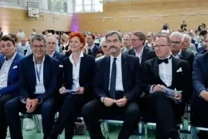 Mehrere Menschen in formeller Kleidung sitzen in einer Sporthalle aufgereiht. Einige von ihnen halten Programme in den Händen. Im Hintergrund sind weitere Personen in der Halle zu sehen.