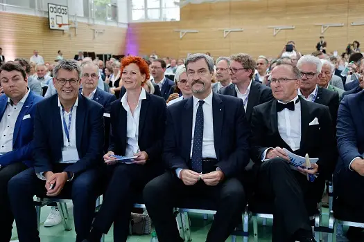 Mehrere Menschen in formeller Kleidung sitzen in einer Sporthalle aufgereiht. Einige von ihnen halten Programme in den Händen. Im Hintergrund sind weitere Personen in der Halle zu sehen.
