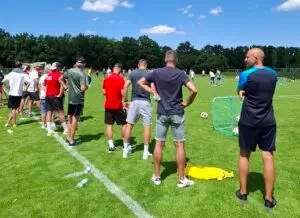 Gruppe von Menschen beobachtet ein Fußballtraining auf einem Rasenplatz. Personen stehen seitlich zum Betrachter und blicken in Richtung Spielfeld, auf dem Spieler mit Bällen trainieren. Bäume sind im Hintergrund sichtbar, der Himmel ist blau mit einigen Wolken.