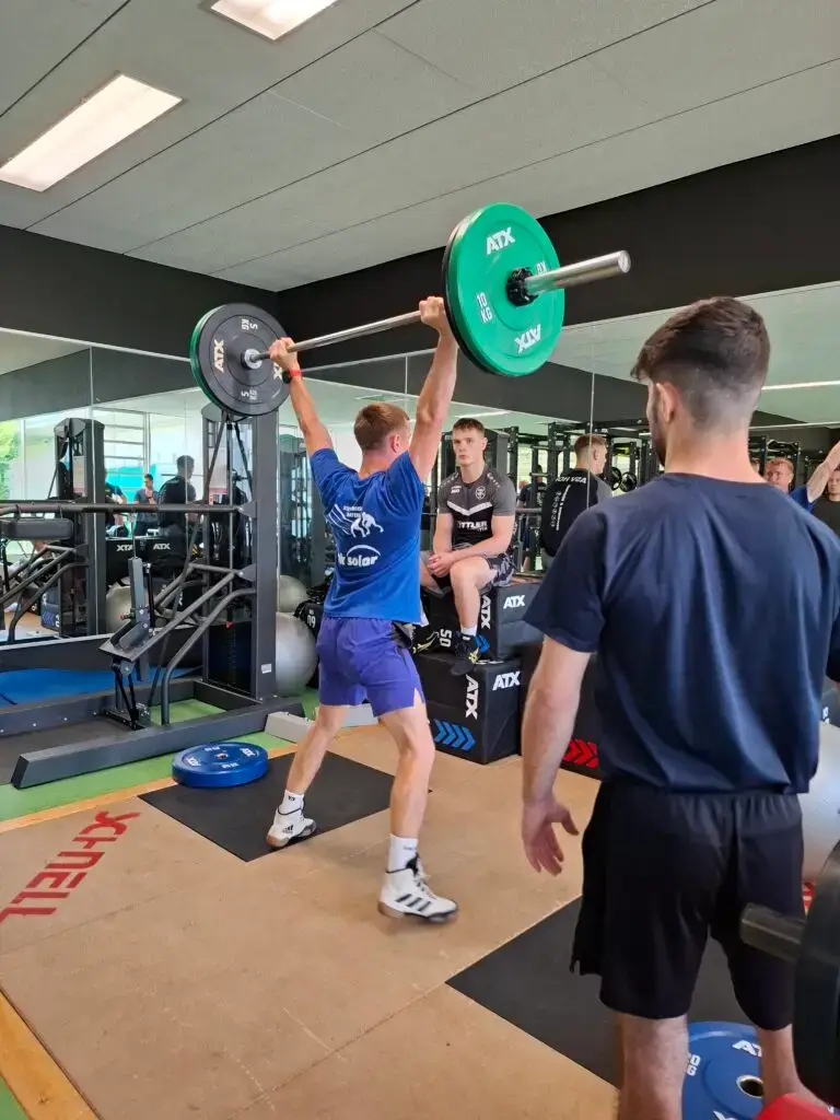 Ein Athlet hebt eine Langhantel über den Kopf in einem Fitnessstudio, während mehrere Personen ihm zuschauen. Im Hintergrund sind Spiegel, Fitnessgeräte und eine Person, die auf einer Box sitzt. Der Raum hat helle Deckenleuchten.