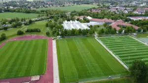 Luftaufnahme eines Sportkomplexes mit mehreren Fu&szlig;ballfeldern, einer Laufbahn und Geb&auml;uden in einer gr&uuml;nen Landschaft