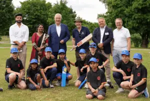 Eine Gruppe von Erwachsenen und Kindern steht zusammen auf einer Wiese. Im Vordergrund knien mehrere Kinder in Sportkleidung mit Baseballcaps, ein Junge hält einen blauen Cricket-Schläger. Hinter ihnen stehen fünf Erwachsene, einer hält einen hölzernen Cricket-Schläger. Die Umgebung ist grün und von Bäumen umgeben.