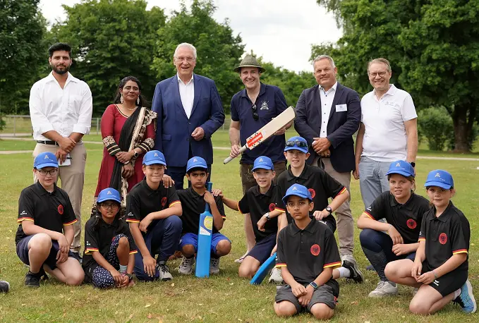 Eine Gruppe von Erwachsenen und Kindern steht zusammen auf einer Wiese. Im Vordergrund knien mehrere Kinder in Sportkleidung mit Baseballcaps, ein Junge hält einen blauen Cricket-Schläger. Hinter ihnen stehen fünf Erwachsene, einer hält einen hölzernen Cricket-Schläger. Die Umgebung ist grün und von Bäumen umgeben.