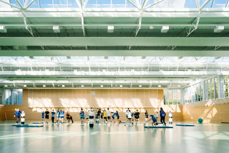 Eine Gruppe von Menschen trainiert in einer hellen Sporthalle mit hohen Decken und großen Fenstern. Der Boden ist mit Turnmatten und Sportgeräten ausgestattet. Lichtstrahlen durchfluten den Raum und werfen Schatten auf die Wände.