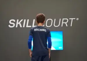 Ein Kind in einem dunklen Sportoutfit mit dem Schriftzug "HACHING" auf dem Rücken steht vor einem Bildschirm, eingebettet in eine Wand mit dem großgeschriebenen Wort "SKILLCOURT".