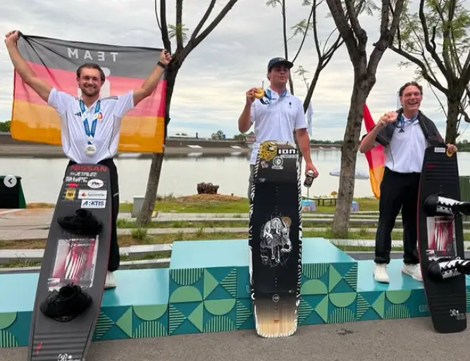Drei Männer stehen auf einem Siegerpodest mit Wakeboards vor sich. Der Mann links hält eine deutsche Flagge und trägt zwei Medaillen, der mittlere hebt eine Goldmedaille hoch und trägt eine silberne Medaille, der rechte hält ebenfalls eine Medaille in der Hand. Sie lächeln und tragen weiße Hemden und dunkle Hosen. Im Hintergrund sind ein See und Bäume zu sehen.
