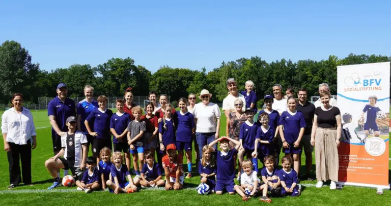 Eine Gruppe von Menschen, darunter Erwachsene und Kinder in blauen Trikots, posiert auf einem Fußballfeld neben einem Werbebanner der BFV Sozialstiftung. Der Himmel ist klar und sonnig, Bäume im Hintergrund.