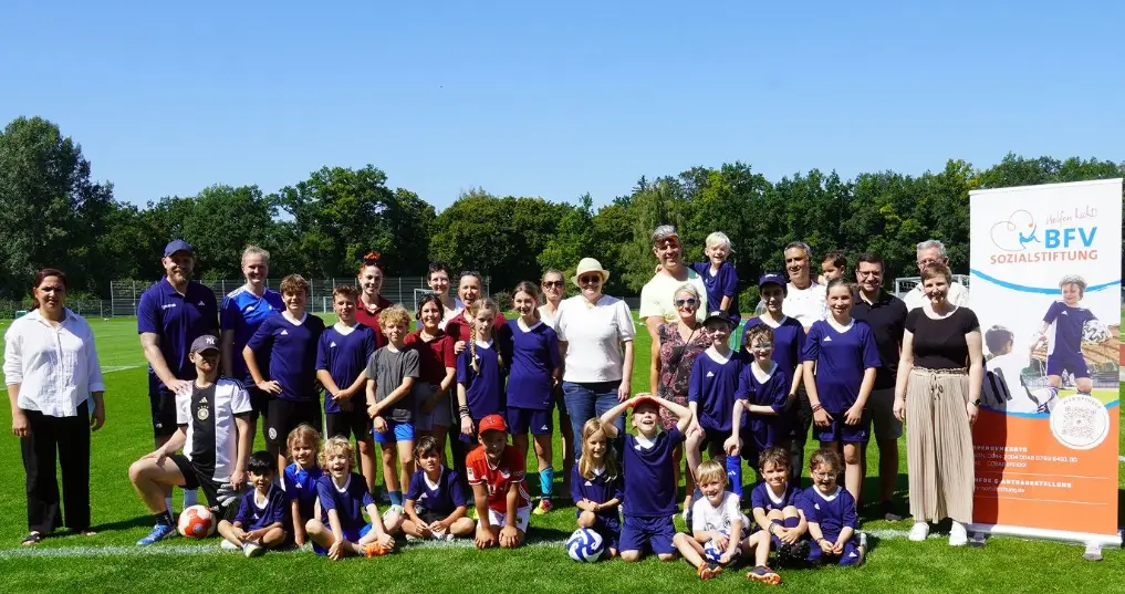 Eine Gruppe von Menschen, darunter Erwachsene und Kinder in blauen Trikots, posiert auf einem Fußballfeld neben einem Werbebanner der BFV Sozialstiftung. Der Himmel ist klar und sonnig, Bäume im Hintergrund.