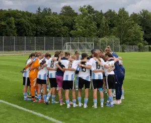 Eine Frauenfußballmannschaft steht gemeinsam im Kreis auf einem Spielfeld, umarmt sich und scheint eine Teambesprechung abzuhalten. Die Spielerinnen tragen einheitliche Trikots in Weiß und Hellblau mit schwarzen Shorts. Im Hintergrund sind ein Tor und ein Zaun zu sehen.