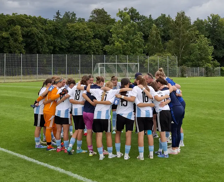 Eine Frauenfußballmannschaft steht gemeinsam im Kreis auf einem Spielfeld, umarmt sich und scheint eine Teambesprechung abzuhalten. Die Spielerinnen tragen einheitliche Trikots in Weiß und Hellblau mit schwarzen Shorts. Im Hintergrund sind ein Tor und ein Zaun zu sehen.