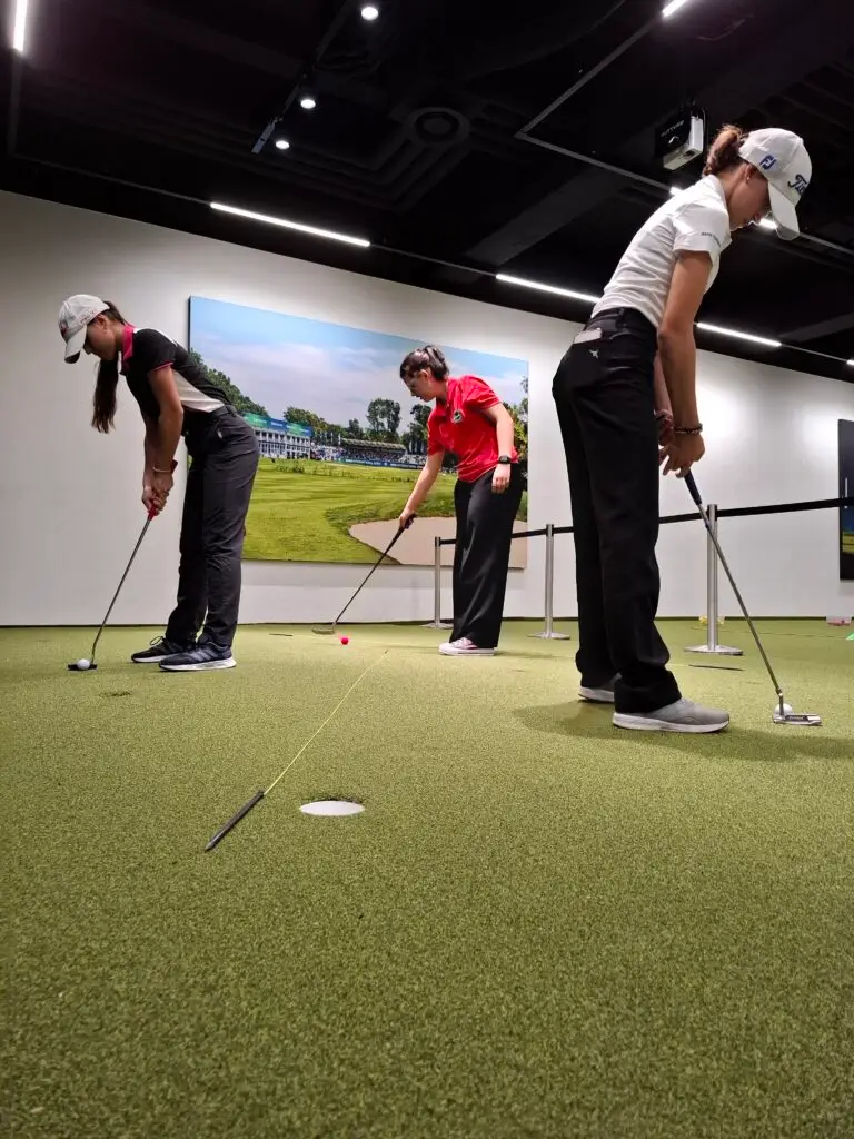 Drei Personen in Sportkleidung praktizieren das Putten auf einem Indoor-Golfgrün. Sie stehen in einer Reihe und konzentrieren sich auf das Einlochen eines Golfballs. Im Hintergrund hängt ein großes Bild eines Golfplatzes an der Wand.