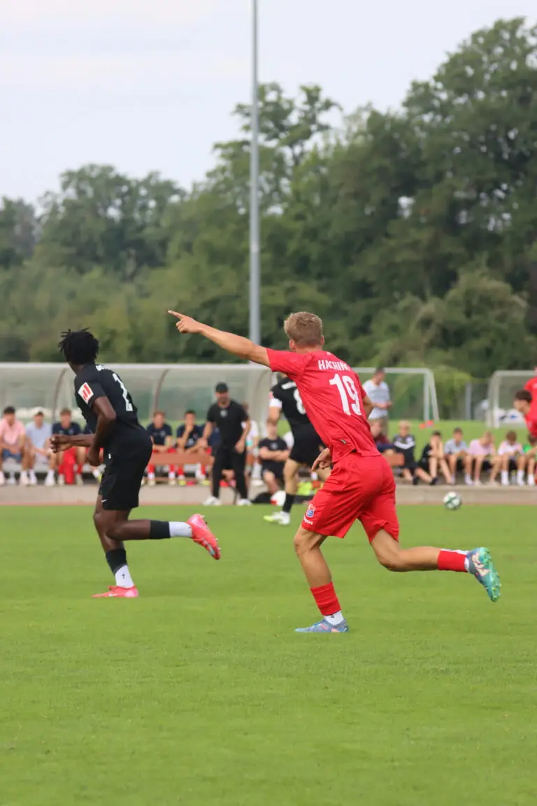 Ein Fußballspiel auf einem Rasenplatz; ein Spieler in einem roten Trikot zeigt mit dem Arm nach vorne, während ein Spieler in einem schwarzen Trikot neben ihm läuft. Im Hintergrund sitzen Zuschauer auf einer Bank am Spielfeldrand.