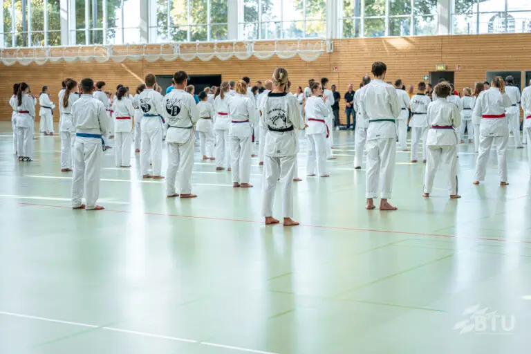 Mehrere Personen im weißen Taekwondo-Anzug stehen in einer Sporthalle. Einige tragen Gürtel in verschiedenen Farben. Im Hintergrund sind Zuschauende zu sehen. Die Halle hat große Fenster, durch die Tageslicht strömt.