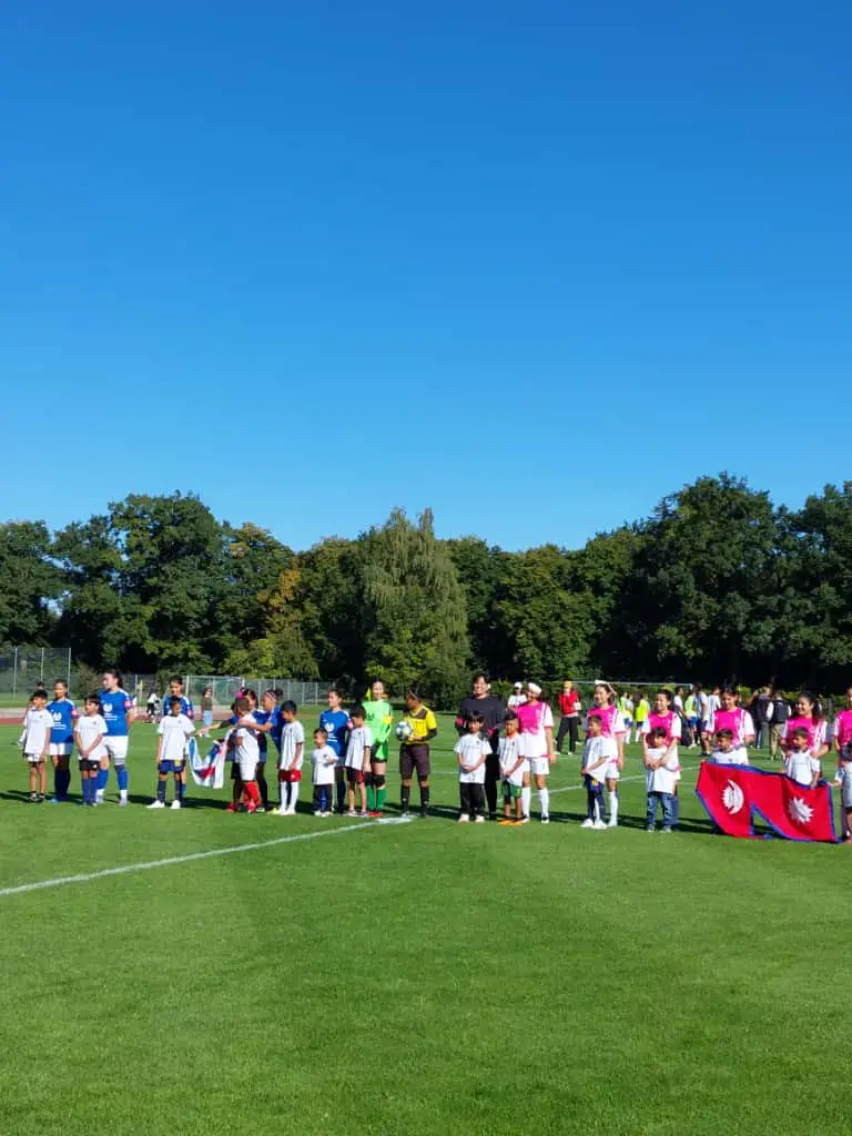 Eine Gruppe von Fußballspielern in blauen und roten Trikots steht auf einem Spielfeld. Kinder in weißen Trikots mit roten und blauen Akzenten stehen vorne. Zwei Kinder halten eine rote Fahne mit weißem Logo. Im Hintergrund sind Bäume und ein klarer, blauer Himmel sichtbar.