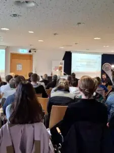 Teilnehmer einer Konferenz sitzen in einem Raum, während eine Frau an einem Rednerpult spricht. Auf der Leinwand ist der Text "7. Bayerische Kinder- und Jugendsportkonferenz" zu lesen. Die Teilnehmer blicken nach vorne zur Präsentation.