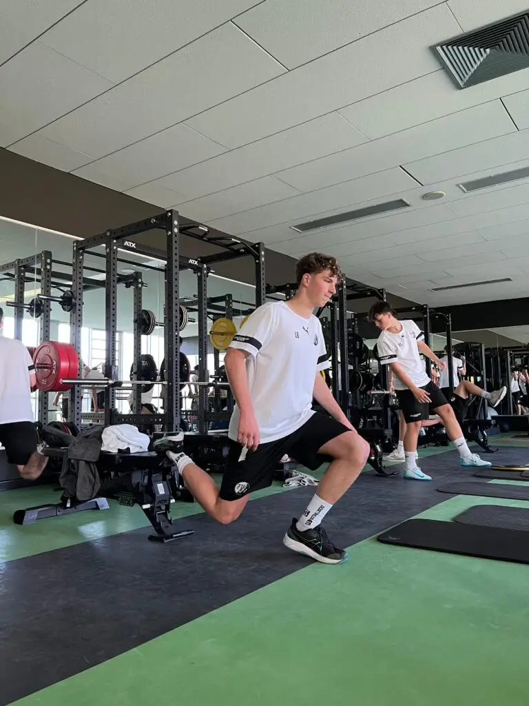 Junge Männer in Sportkleidung führen Kniebeugen in einem Fitnessstudio durch. Sie nutzen Bänke zur Unterstützung, während im Hintergrund freie Gewichte und Spiegel sichtbar sind.