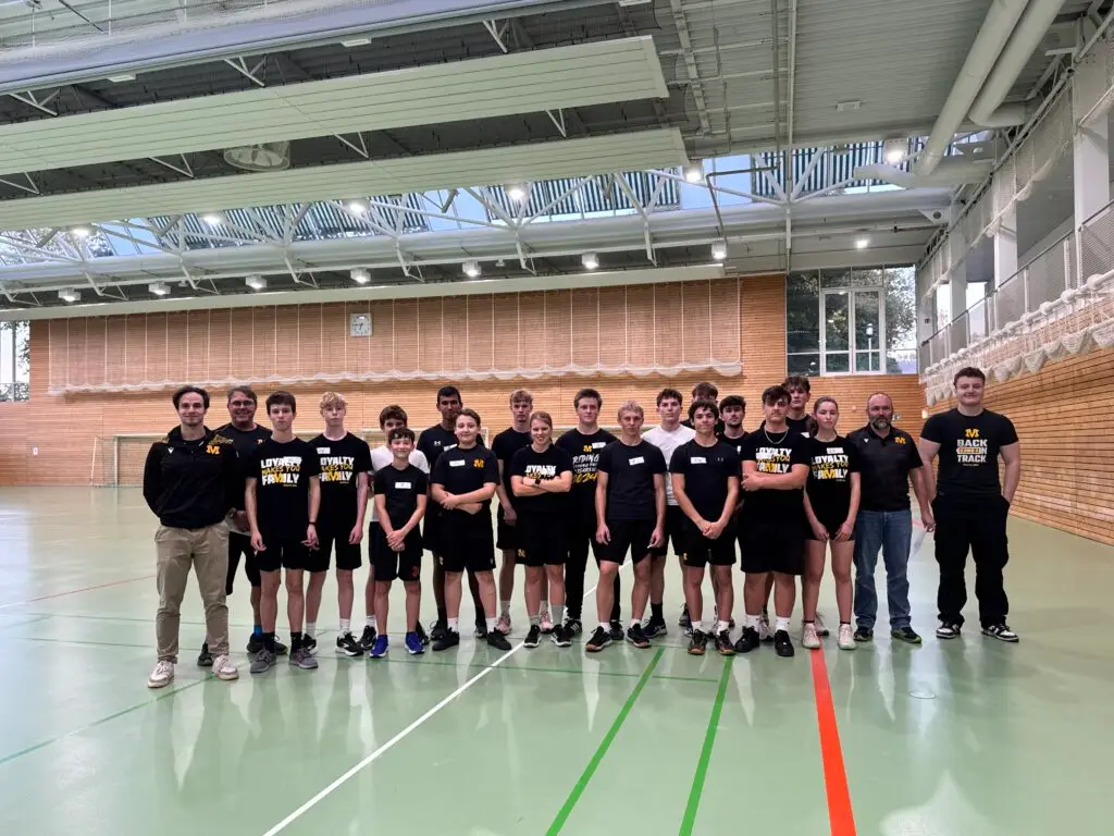 Gruppe von vierzehn Teenagern und vier Erwachsenen, alle in Sportkleidung, steht in einer Turnhalle mit Holzwänden und großen Fenstern. Die Jugendlichen tragen passende Shirts, einige mit dem Aufdruck "LOYALTY FAMILY". Zwei Erwachsene stehen links, zwei weitere rechts am Rand. Der Boden ist mit Linien für verschiedene Sportarten markiert.