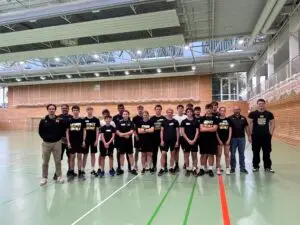 Gruppe von vierzehn Teenagern und vier Erwachsenen, alle in Sportkleidung, steht in einer Turnhalle mit Holzwänden und großen Fenstern. Die Jugendlichen tragen passende Shirts, einige mit dem Aufdruck "LOYALTY FAMILY". Zwei Erwachsene stehen links, zwei weitere rechts am Rand. Der Boden ist mit Linien für verschiedene Sportarten markiert.