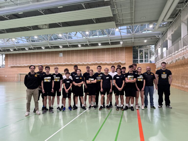 Eine Gruppe von jungen Sportlern und Trainern steht in einer Sporthalle. Die meisten tragen schwarze Sportkleidung mit gelbem Logo. Im Hintergrund eine große, helle Sporthalle mit hohen Decken und einer Uhr an der Wand.