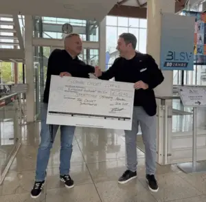 Zwei Männer stehen in einem Gebäude und lächeln sich an, während sie sich die Hand geben. Sie halten zusammen einen überdimensionalen Scheck mit der Aufschrift "German Cricket Club e.V." Der Scheck ist auf 1210 Euro ausgestellt. Im Hintergrund sind Treppen, große Fenster und ein Schild mit der Aufschrift "BLSV 80 Jahre" sichtbar.