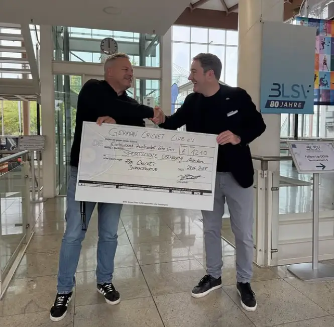 Zwei Männer stehen in einem Gebäude und lächeln sich an, während sie sich die Hand geben. Sie halten zusammen einen überdimensionalen Scheck mit der Aufschrift "German Cricket Club e.V." Der Scheck ist auf 1210 Euro ausgestellt. Im Hintergrund sind Treppen, große Fenster und ein Schild mit der Aufschrift "BLSV 80 Jahre" sichtbar.