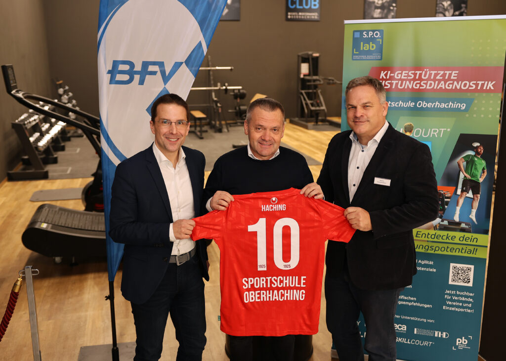 Auf dem Foto stehen drei Männer in einem Trainings- und Diagnostikbereich der Sportschule Oberhaching. Links ist BFV-Präsident Dr. Christoph Kern, in der Mitte SpVgg Unterhaching-Präsident Manfred Schwabl und rechts SPO-Geschäftsführer Markus Mayer zu sehen. Gemeinsam halten sie ein rotes Trikot mit der Aufschrift „Haching“ sowie „Sportschule Oberhaching“ und der Rückennummer 10 in die Kamera. Im Hintergrund sind Trainingsgeräte, eine BFV-Fahne sowie ein S.P.O. lab-Roll-up mit dem Thema „KI-gestützte Leistungsdiagnostik“ erkennbar.