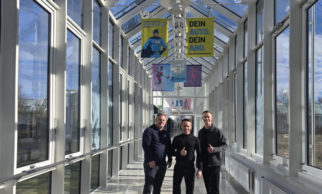 Drei Personen stehen in einem hellen, verglasten Gang der Sportschule Oberhaching und blicken in die Kamera; über ihnen hängen Banner, im Hintergrund sind Sportmotive und Außenbereiche sichtbar.
