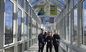Drei Personen stehen in einem hellen, verglasten Gang der Sportschule Oberhaching und blicken in die Kamera; über ihnen hängen Banner, im Hintergrund sind Sportmotive und Außenbereiche sichtbar.