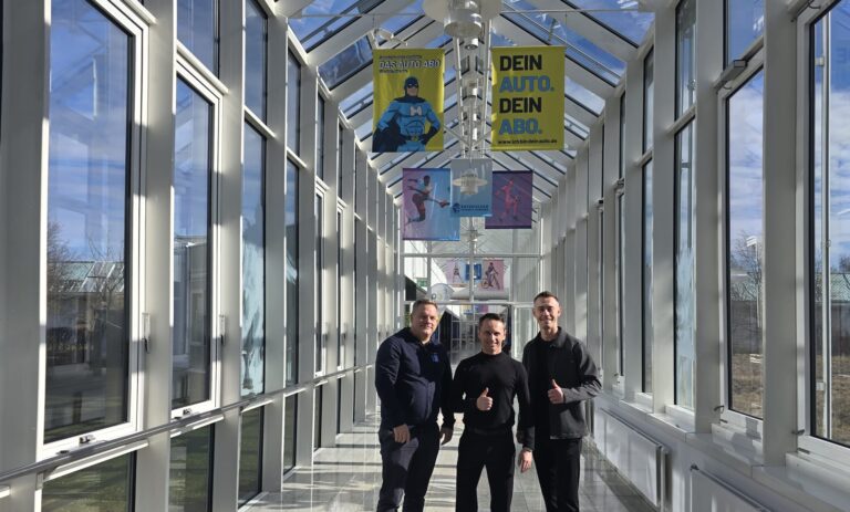 Drei Personen stehen in einem hellen, verglasten Gang der Sportschule Oberhaching und blicken in die Kamera; über ihnen hängen Banner, im Hintergrund sind Sportmotive und Außenbereiche sichtbar.