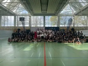 Gruppenfoto des 17. Kids Camps der Peak Performer Stiftung in einer großen, lichtdurchfluteten Sporthalle der Sportschule Oberhaching: Rund 100 Kinder und Betreuer sitzen und stehen gemeinsam auf dem Hallenboden vor einem Banner, umgeben von großen Fensterfronten und Sportfeldmarkierungen.