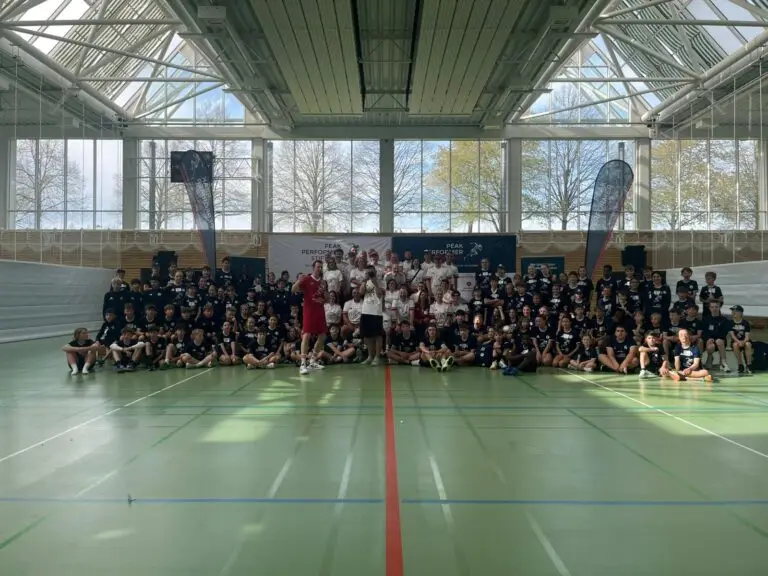 Gruppenfoto des 17. Kids Camps der Peak Performer Stiftung in einer großen, lichtdurchfluteten Sporthalle der Sportschule Oberhaching: Rund 100 Kinder und Betreuer sitzen und stehen gemeinsam auf dem Hallenboden vor einem Banner, umgeben von großen Fensterfronten und Sportfeldmarkierungen.