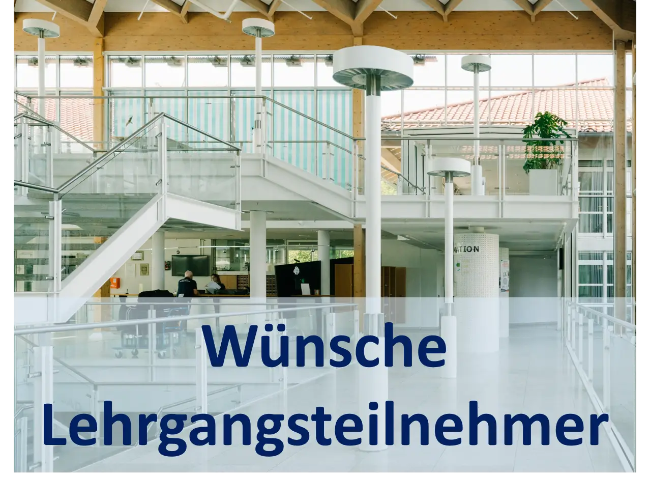 Heller Innenbereich eines modernen Geb&auml;udes mit Glasgel&auml;ndern, Treppen und Holzdecken; im Hintergrund ein Empfangsbereich, dar&uuml;ber ein halbtransparenter Banner mit dem Text &bdquo;W&uuml;nsche Lehrgangsteilnehmer&ldquo;.