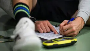 Nahaufnahme einer Leistungsdiagnostik: Eine Person tr&auml;gt Messsensoren am Bein, w&auml;hrend Testergebnisse auf einem Dokument notiert werden. Im Vordergrund liegt ein mobiles Messger&auml;t auf einem Tisch.