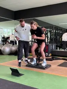 Basketballspielerin absolviert im Kraftraum der Sportschule Oberhaching eine leistungsdiagnostische Schnellkraftmessung auf einer Messplattform, begleitet und angeleitet von einem Athletiktrainer.