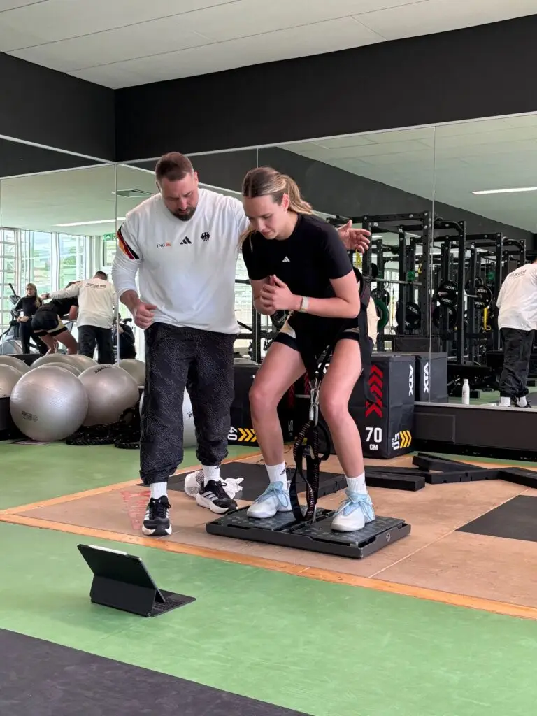 Basketballspielerin absolviert im Kraftraum der Sportschule Oberhaching eine leistungsdiagnostische Schnellkraftmessung auf einer Messplattform, begleitet und angeleitet von einem Athletiktrainer.
