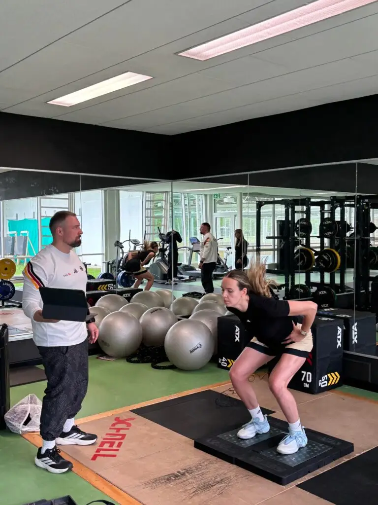 Basketballspielerin f&uuml;hrt im Kraftraum der Sportschule Oberhaching einen Sprung- beziehungsweise Schnellkrafttest auf einer Messplattform durch, w&auml;hrend ein Athletiktrainer die Leistungsdiagnostik mit einem Tablet begleitet.