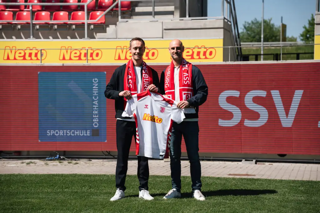 Alexander Schmalhofer, Sportdirektor des SSV Jahn und Niklas Reuter (Leitung Marketing & Projektmanagement der Sportschule Oberhaching) beim gemeinsamen Foto. Beide halten ein Trikot des SSV Jahn Regensburg hoch.