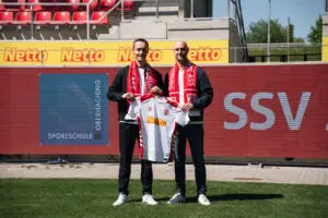 Alexander Schmalhofer, Sportdirektor des SSV Jahn und Niklas Reuter (Leitung Marketing & Projektmanagement der Sportschule Oberhaching) beim gemeinsamen Foto. Beide halten ein Trikot des SSV Jahn Regensburg hoch.