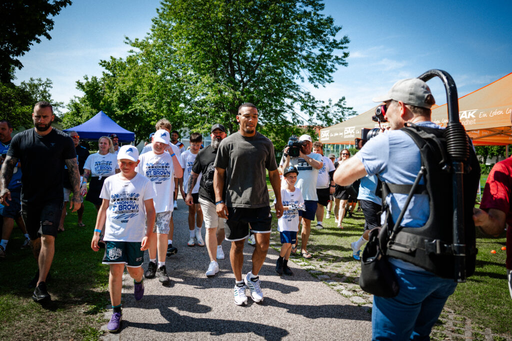 Kinder und Jugendliche begleiten NFL Star Amon-Ra St. Brown während eines American-Football-Camps auf dem Gelände der Sportschule Oberhaching.“