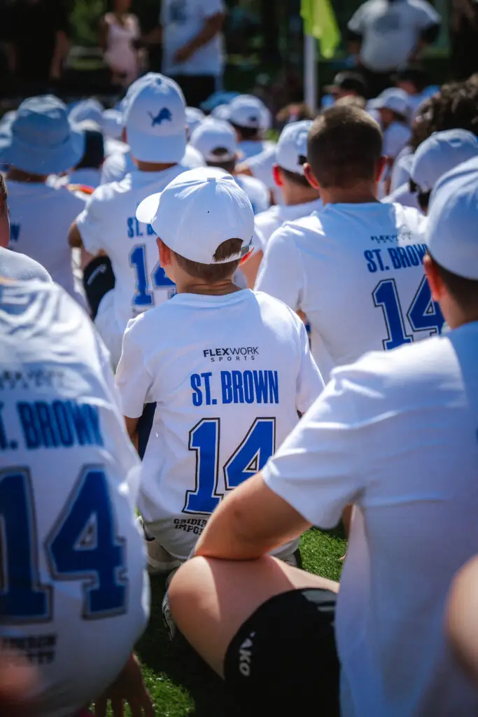 Kinder und Jugendliche sitzen während eines American-Football-Camps auf dem Kunstrasenplatz und tragen weiße Camp-Shirts sowie Caps.