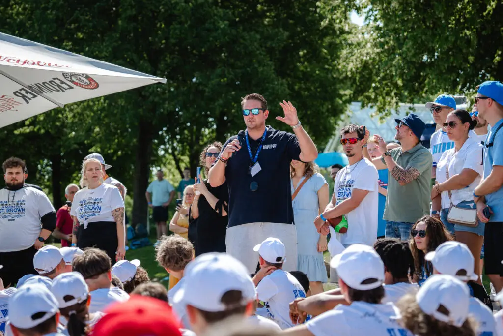 Organisator begrüßt die Teilnehmenden eines Football-Camps vor einer großen Gruppe von Kindern, Jugendlichen und Begleitpersonen