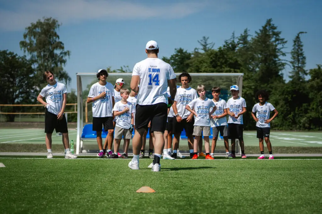 rainer erklärt einer Gruppe von Kindern und Jugendlichen die nächste Übung während eines American-Football-Camps auf dem Kunstrasenplatz.